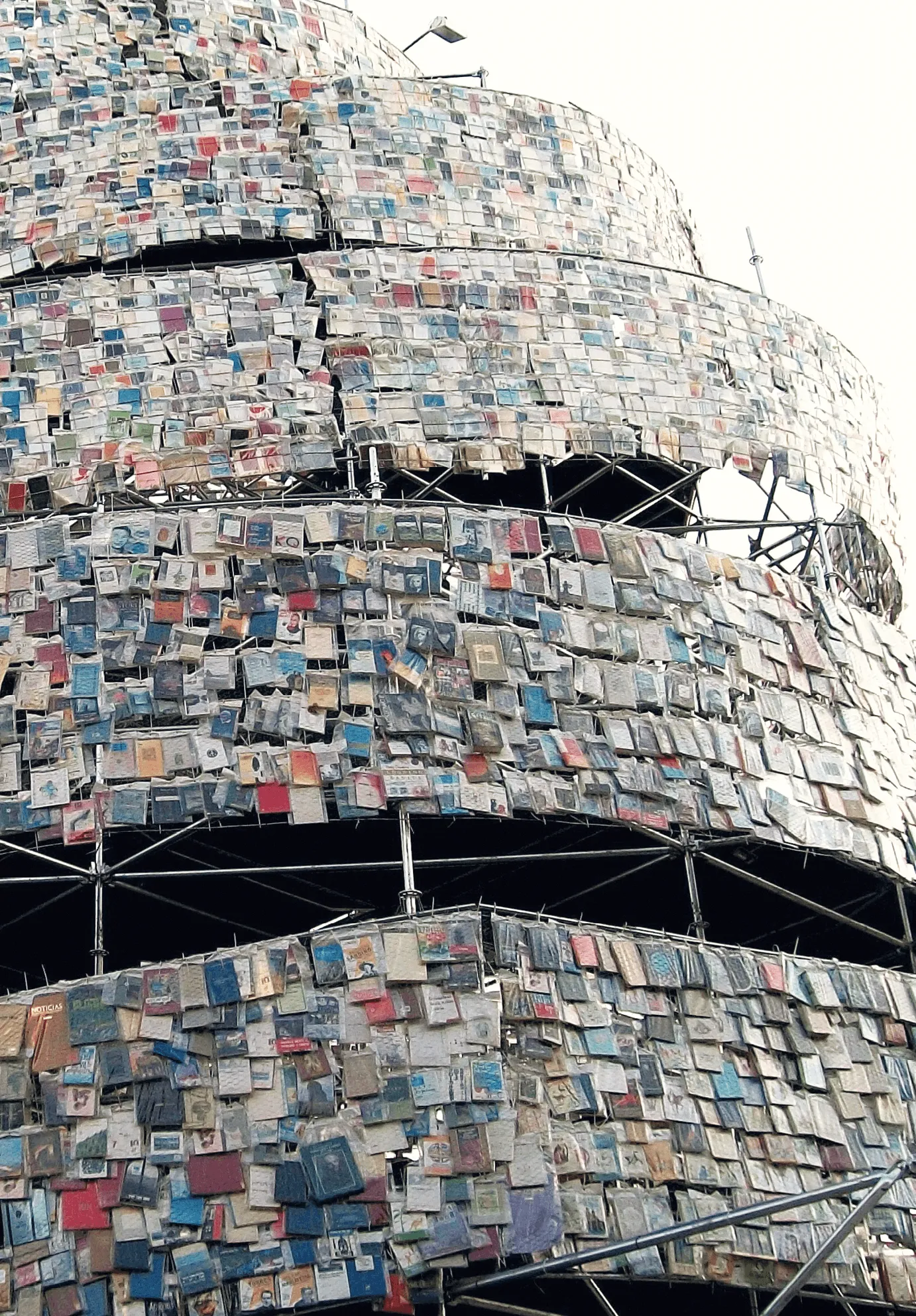 Photographie d'une structure recouverte de livres, évoquant une tour de Babel. Des milliers de livres de différentes couleurs et tailles sont fixés sur une armature métallique.