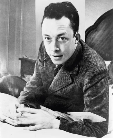 Photographie noir et blanc d'Albert Camus, écrivant, assis sur un lit, vêtu d'un costume à carreaux.