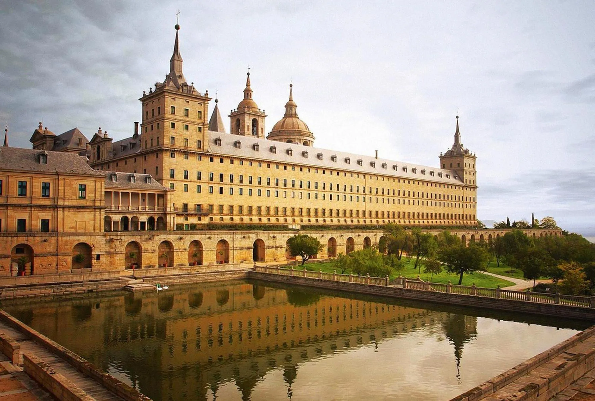 Photographie du monastère royal de San Lorenzo de l'Escurial, Espagne.  Grand bâtiment beige, reflété dans un bassin d'eau. Architecture imposante et jardin.