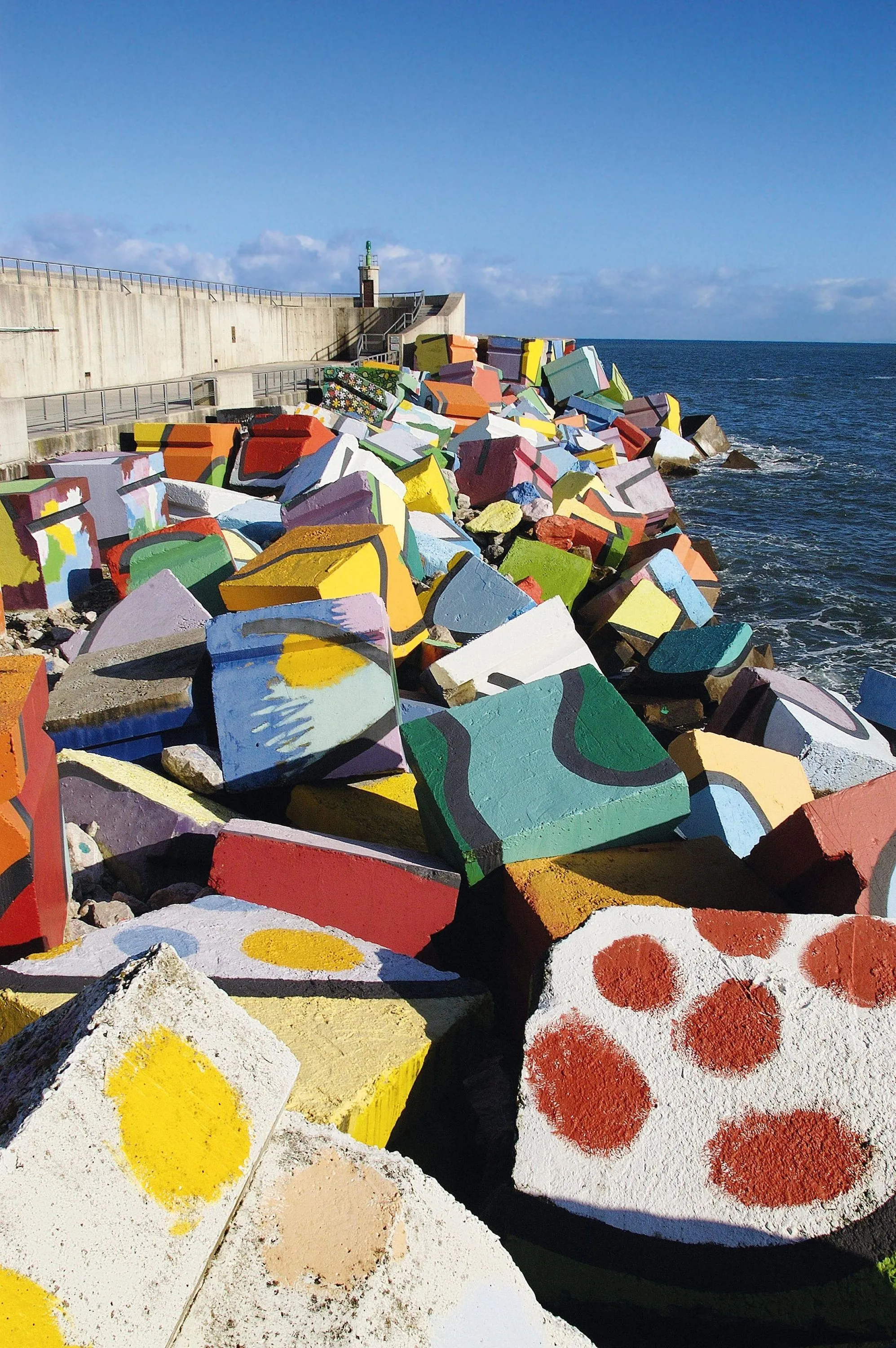 Photographie de blocs de béton colorés disposés sur un brise-lames, près d'un phare. Art abstrait, couleurs vives.