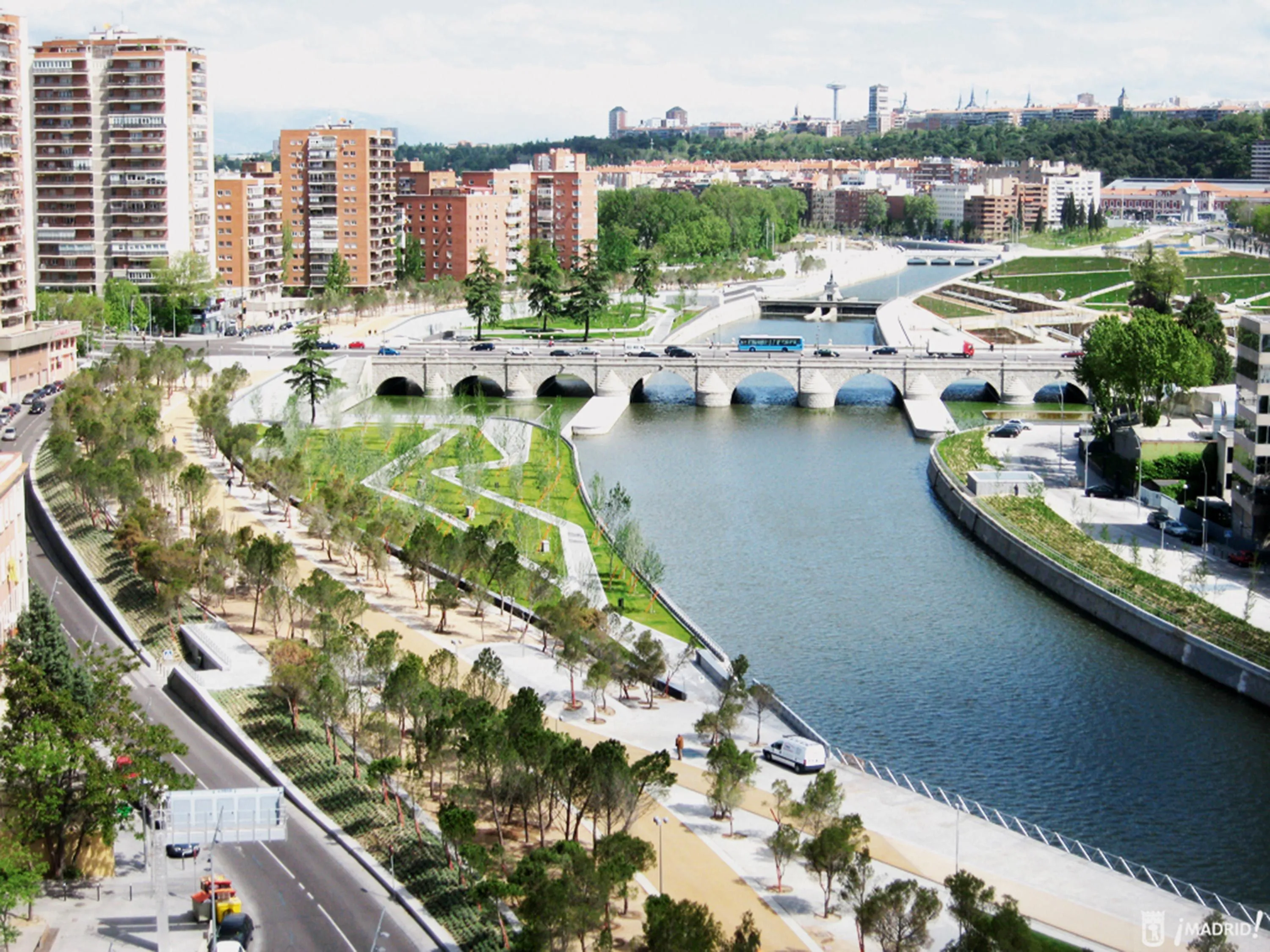 Photographie aérienne d'un parc urbain longeant une rivière à Madrid. Un pont en pierre traverse la rivière. Bâtiments modernes en arrière-plan.