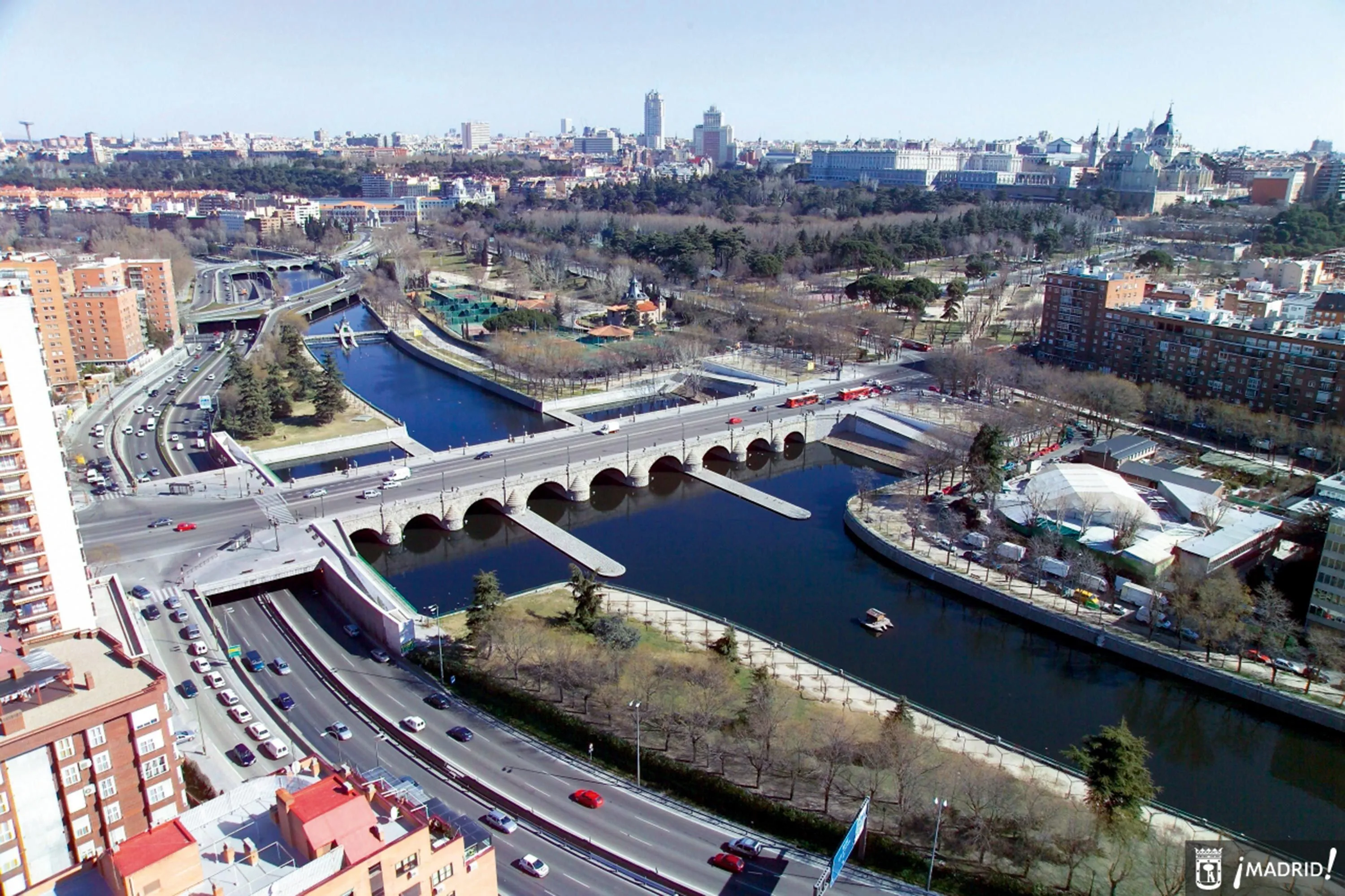 Photo aérienne d'un pont à arches sur une rivière à Madrid, entouré de verdure et de bâtiments. Vue panoramique sur la ville en arrière-plan.