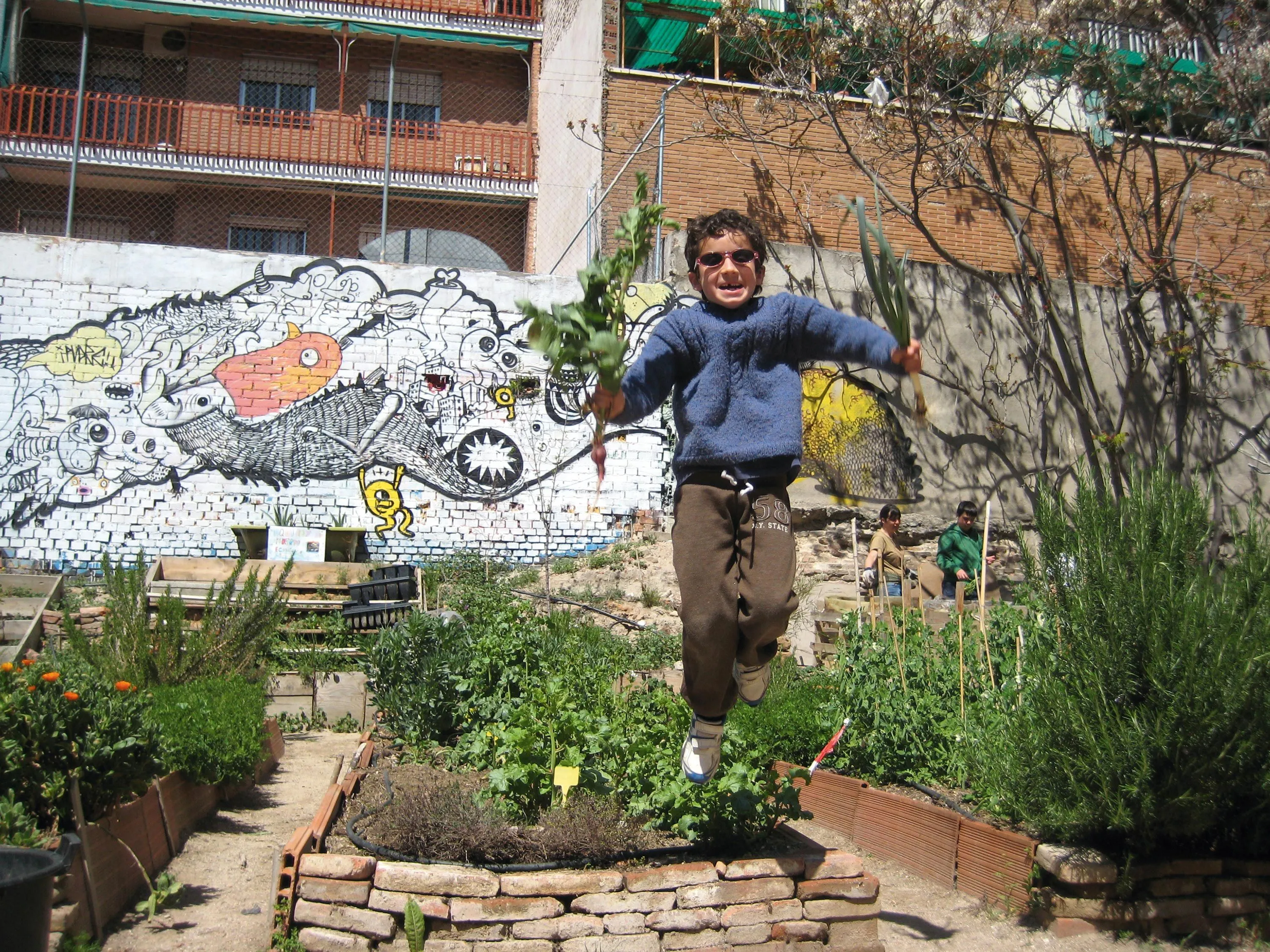 Photographie d'un enfant sautant dans un jardin urbain, tenant des légumes. Mur de graffitis en arrière-plan.