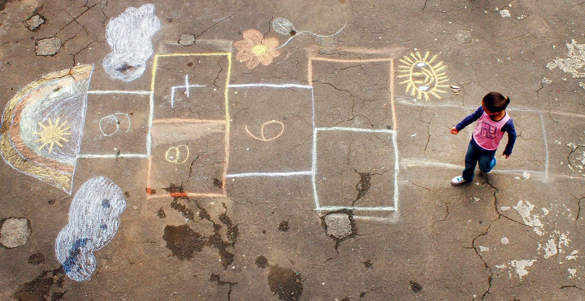 Photographie aérienne d'une fillette jouant à la marelle sur le sol fissuré. Dessins à la craie : arc-en-ciel, soleil, nuages.