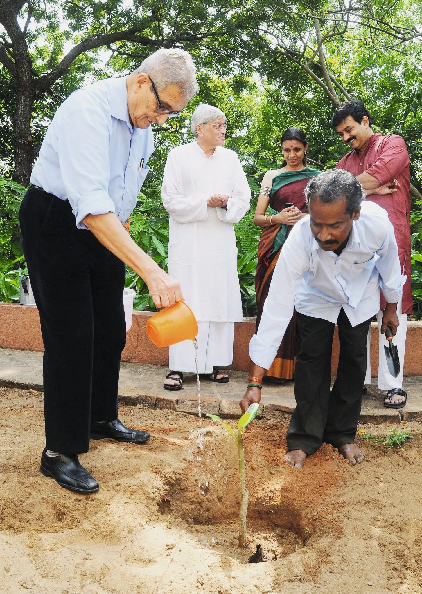 Photographie d'Amartya Sen arrosant un jeune plant à Chennai en Inde, en 2013, assisté par d'autres personnes.