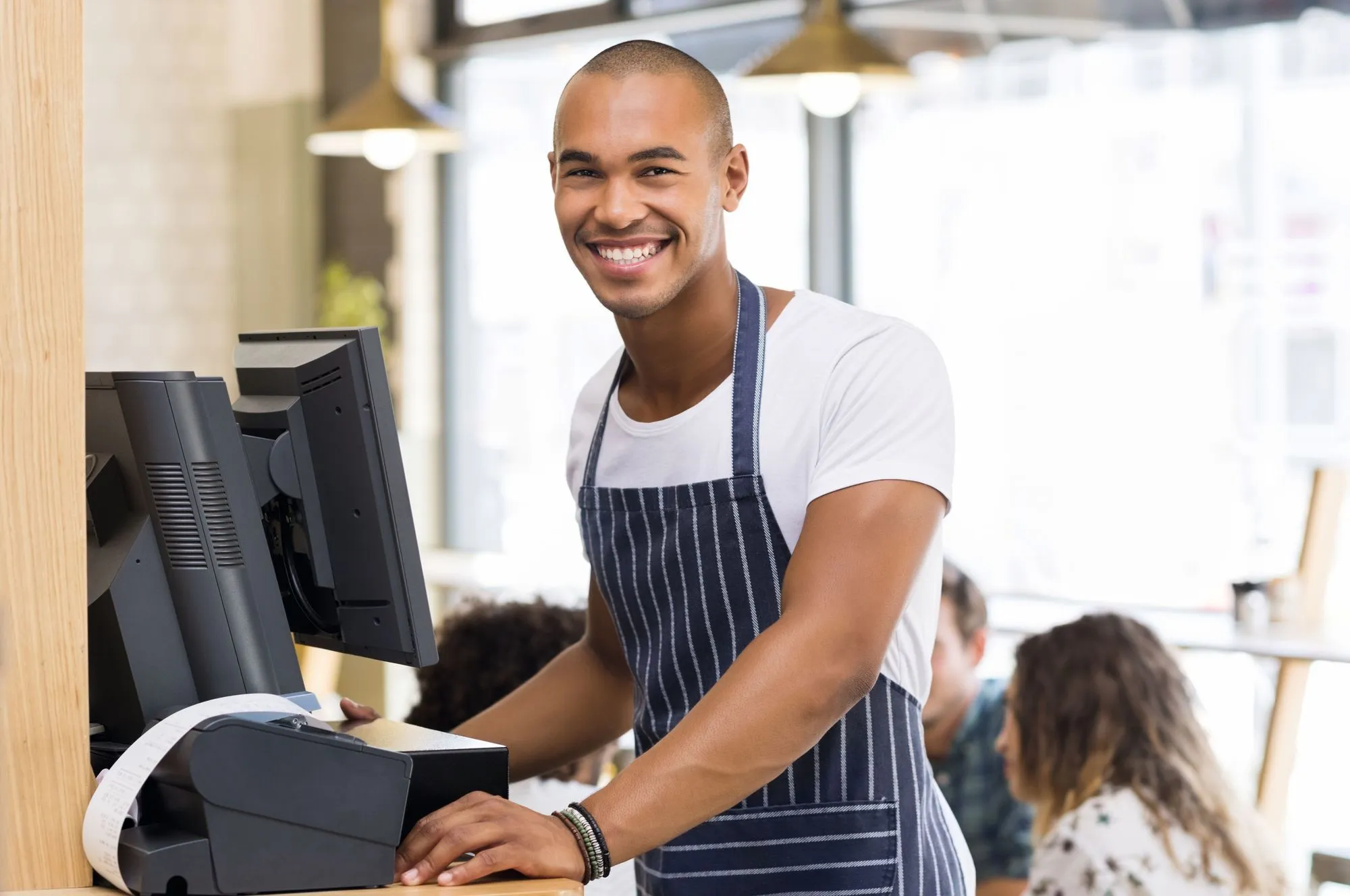 Photographie d'un serveur souriant derrière une caisse enregistreuse dans un café. Il porte un tablier rayé. Clients flous en arrière-plan.