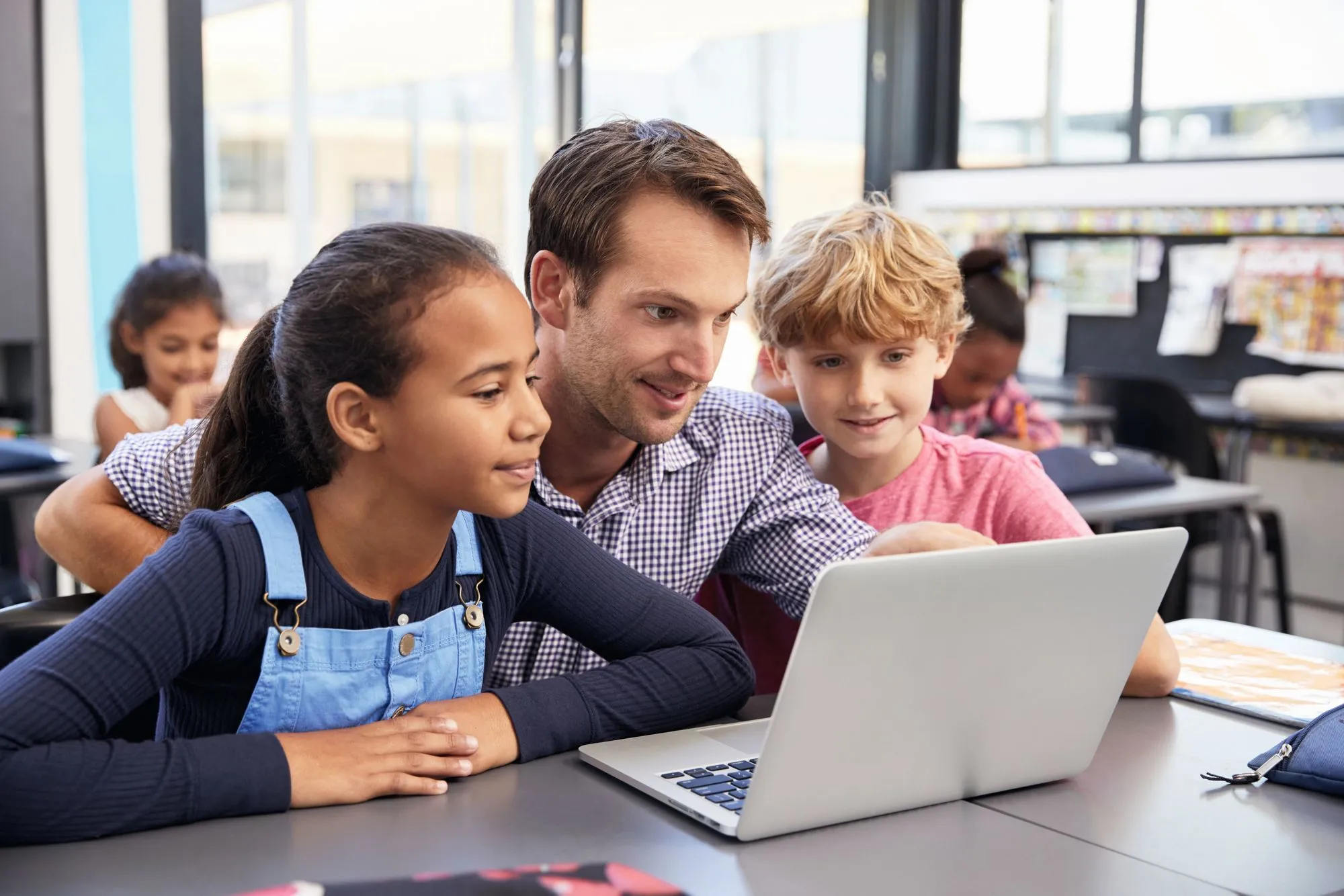 Photographie : un enseignant aide deux élèves à utiliser un ordinateur portable en classe.