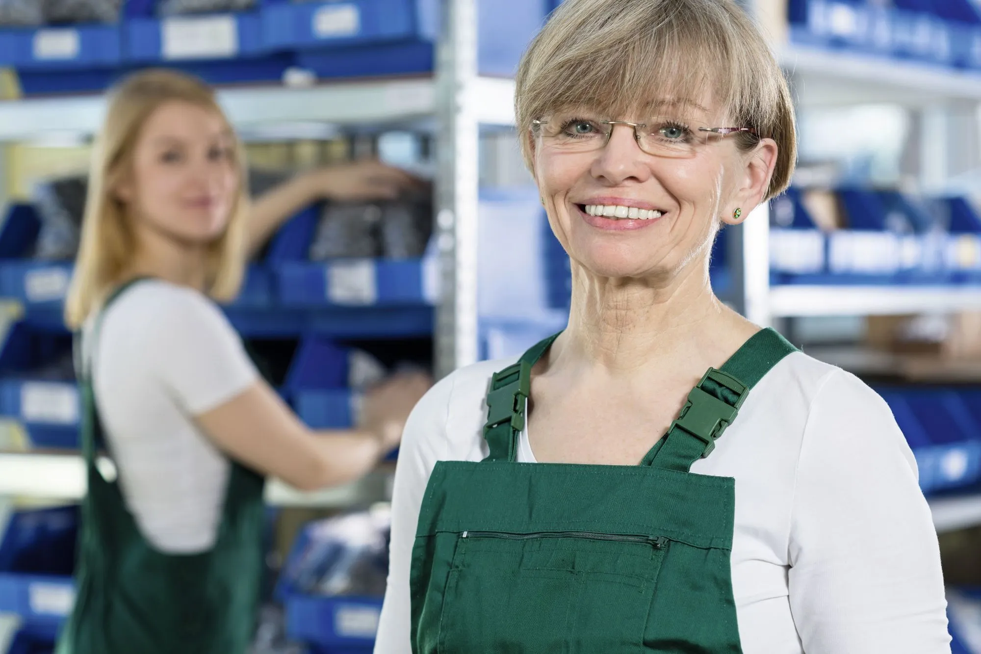 Photographie d'une femme souriante en salopette verte, une autre femme est floue en arrière-plan. Cadre industriel.