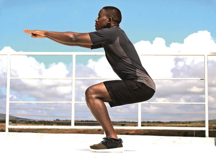 Photographie d'un homme effectuant un squat en extérieur, par temps ensoleillé.  L'homme, vêtu de noir, est face à un ciel bleu et nuageux.