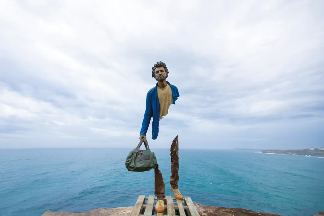 Photographie d'une sculpture en bronze de Bruno Catalano, représentant un homme incomplet tenant un sac, près de l'océan à Sydney.