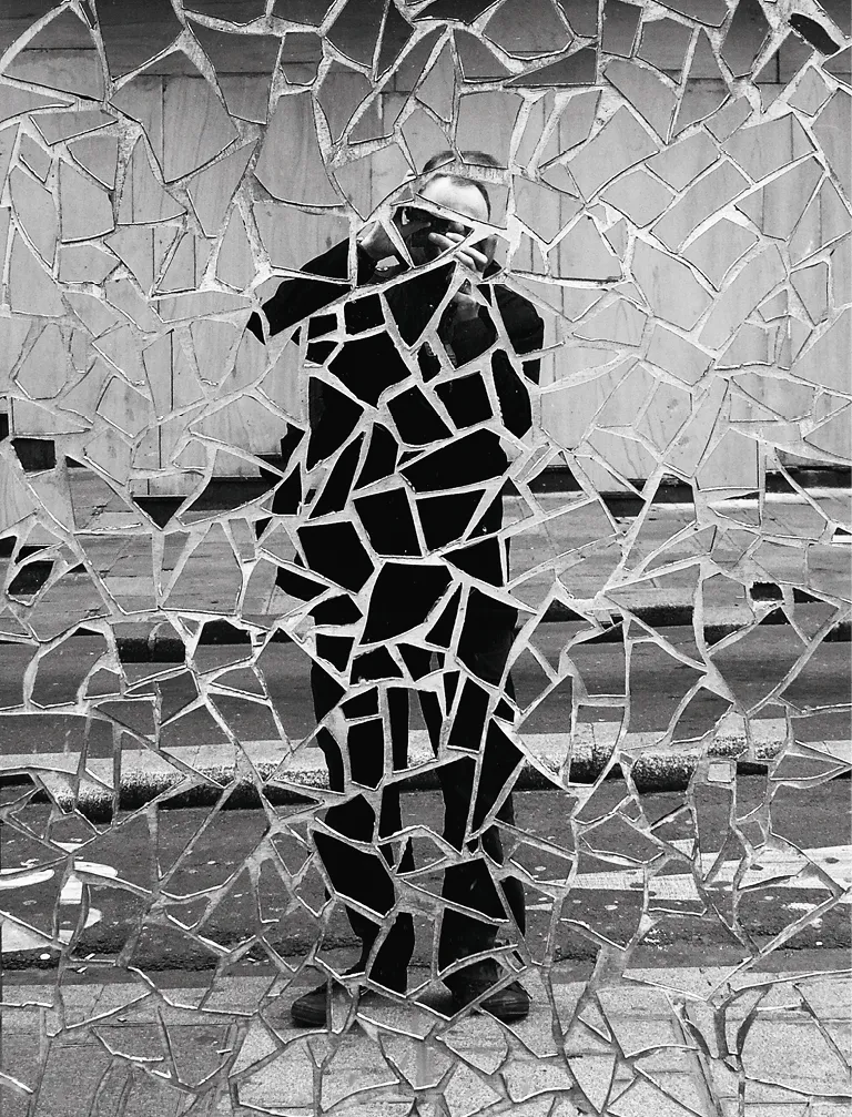 Photographie monochrome d'un autoportrait fragmenté. L'homme est vu à travers un miroir brisé en plusieurs morceaux, créant un effet de mosaïque.