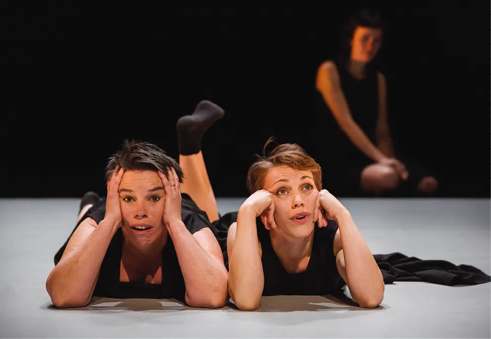 Photographie de scène de théâtre : deux femmes gisant au sol, mains sur le visage, regardent vers le haut, une troisième femme floue en arrière-plan.