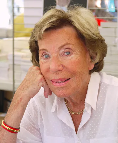 Photographie de Benoîte Groult, écrivaine française, le visage reposant sur la main, souriante, portant un chemisier blanc et un bracelet rouge.